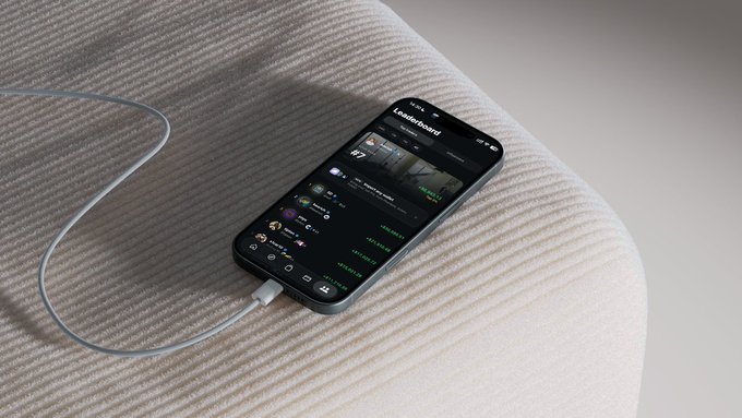 A smartphone rests on a ribbed fabric surface with a charging cable attached, displaying a dark-mode sports scoreboard app titled "Leaderboard" with various team scores and logos.