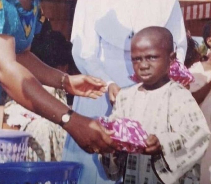 A vintage photograph showing a young child with a serious expression wearing a patterned white garment, being handed a pink wrapped item by an adult wearing white, with a blue basket visible in the foreground.