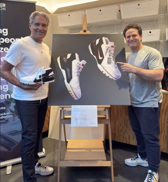 Two men standing on either side of a canvas displaying a custom sneaker design; the man on the left holds the physical shoe while the man on the right points at the artwork.