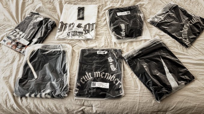 A flat-lay photograph shows several folded black and white graphic t-shirts in clear plastic packaging spread across a wrinkled white bedsheet.