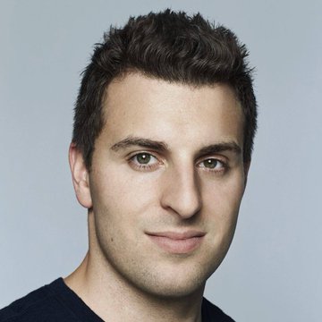 A professional headshot of a man with short dark hair, brown eyes, and slight stubble, wearing a dark t-shirt against a solid light blue background.