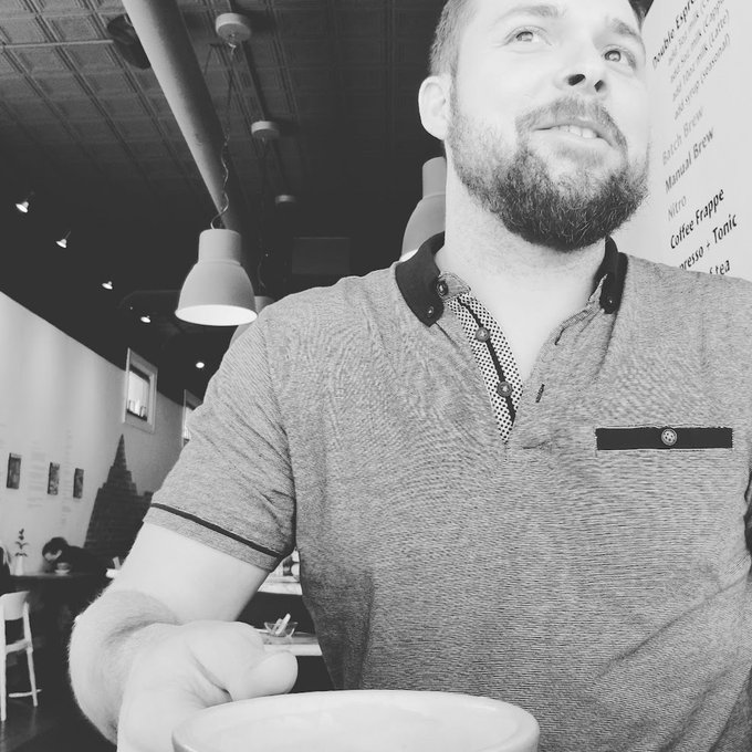A black-and-white selfie of a bearded man wearing a polo shirt, looking upward, with a menu board listing items like 'Nitro Coffee Frappe' visible in the background.
