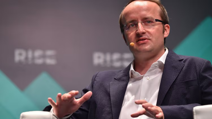 A man wearing glasses and a dark suit speaks at a conference, gesturing with his hands against a backdrop displaying the 'RISE' logo.