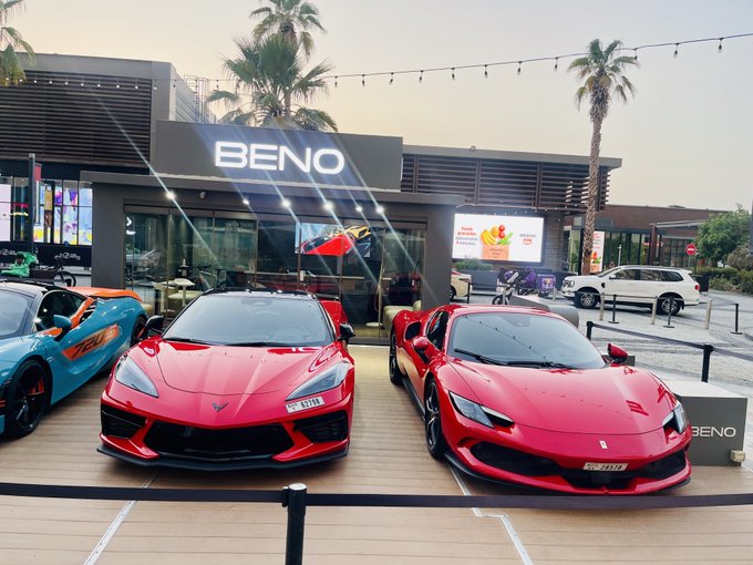 Two red sports cars (appearing to be a Chevrolet Corvette) are parked side by side outside a restaurant labeled "BENO," with a palm tree and modern building facade in the background.