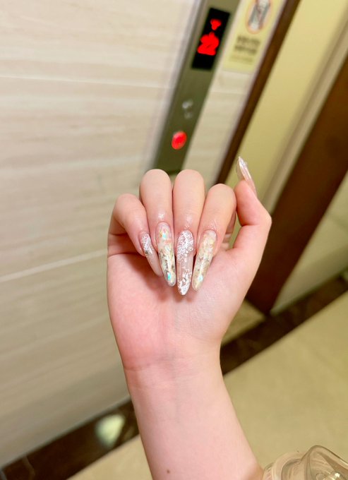 A close-up of a hand displaying long, pointed nails with iridescent glitter and pastel colors in front of an elevator with a red digital floor indicator.