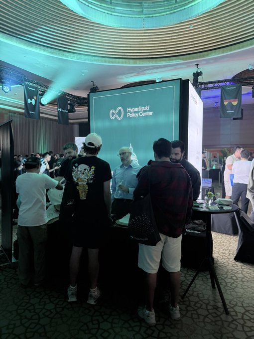 A photo of a crowded conference booth labeled 'Hyperliquid Policy Center' where several attendees in casual attire are gathered around a counter in a venue with a curved ceiling.