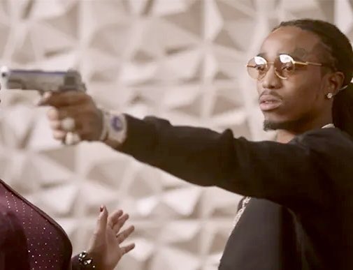 A man wearing round sunglasses and a black shirt pointing a silver handgun, appearing to be a still from a music video.