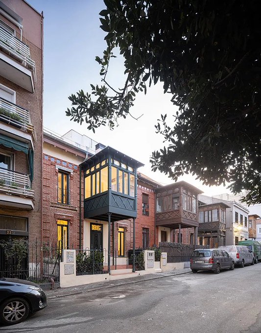 A row of charming townhouses featuring distinctive enclosed glass balconies lines a quiet Madrid street, with the brick-and-stone facades bathed in soft golden light beneath a leafy tree canopy. Parked cars and a gated entrance suggest the upscale residential character of this Castelar neighborhood.