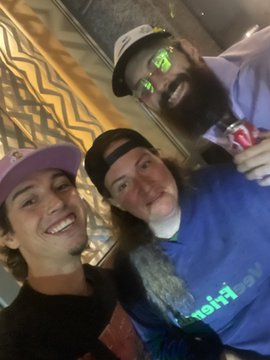 Three men pose together for a close-up selfie at what appears to be a sporting event, with one wearing reflective green sunglasses.
