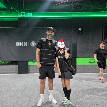 A man and child pose together in a soccer-themed arena with "OKX" branding on the walls, with the child holding a red mascot plushie. The scene reflects the Manchester City Legends Event with OKX, where fans got to meet players like Shaun Wright-Phillips and Scott Carson and play mini-games together.