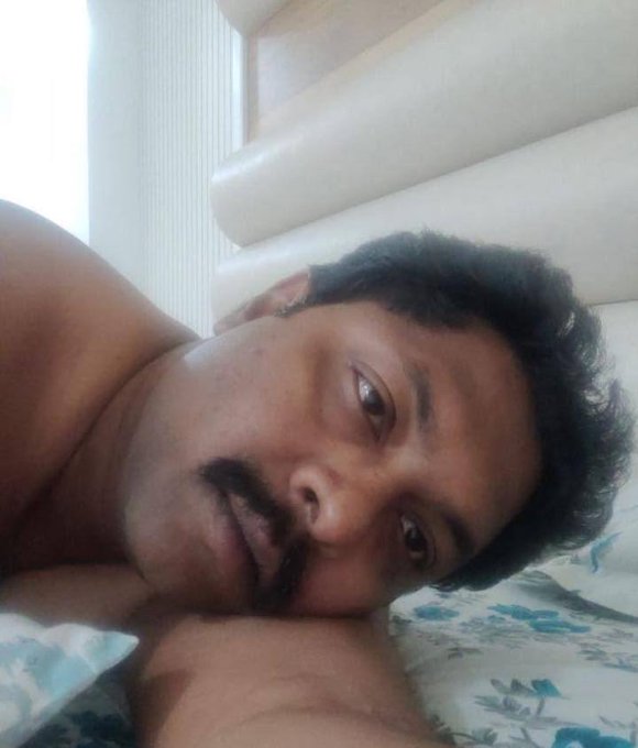 A close-up selfie of a man with a mustache lying down against a white cushion with floral patterned bedding.