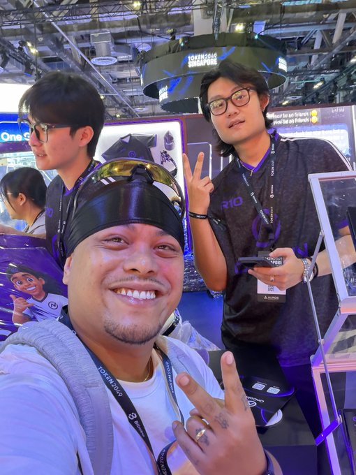 Three people posing at what appears to be a tech convention (Token2049 event), with two making peace signs and wearing lanyards.
