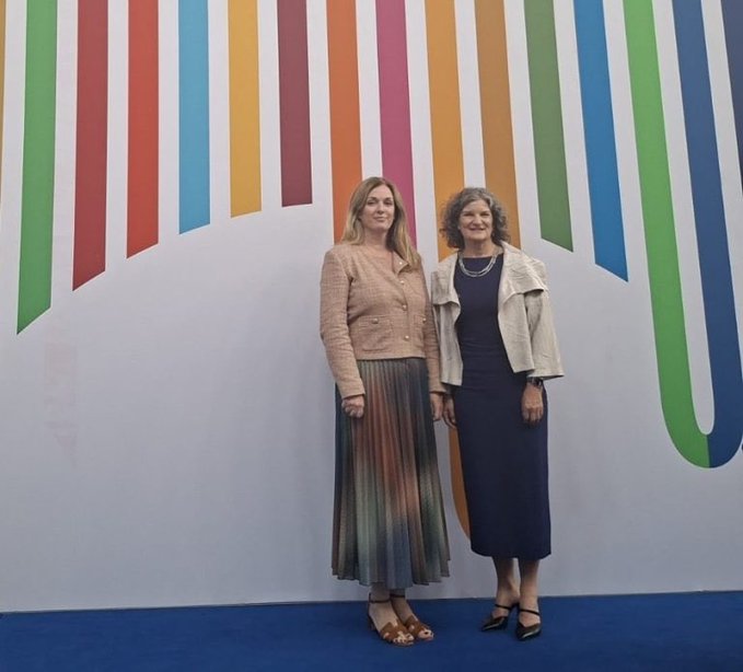Two women stand together posing in front of a colorful striped backdrop featuring a large stylized "UN" logo, representing the partnership between Stellar and UNDP to deliver blockchain-based digital assistance more efficiently during crises.