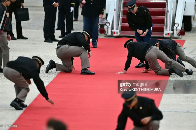 A Getty Images photo by Kent Nishimura showing uniformed staff crawling on their hands and knees to inspect a red carpet before an event at what appears to be an airport with stairs visible.