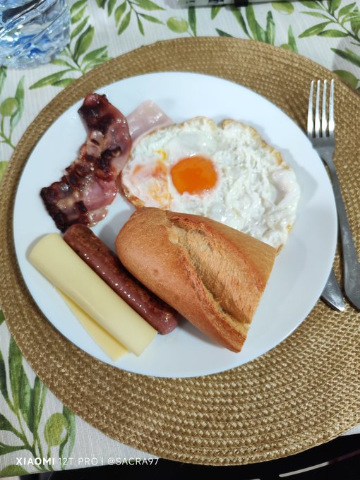 The image shows a plate of food, including eggs, bacon, cheese, bread, and sausage, placed on a table with a woven placemat underneath. There are also utensils like a fork and knife present on the table. The main focus is on the delicious breakfast meal that includes various types of food items arranged neatly on the plate.