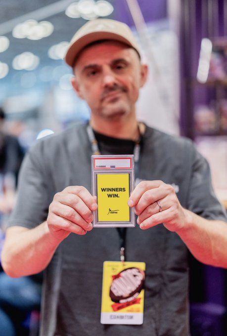 A man wearing a beige cap and gray shirt at what appears to be a convention, holding up a yellow card that reads 'WINNERS WIN.' with an exhibitor badge around his neck.