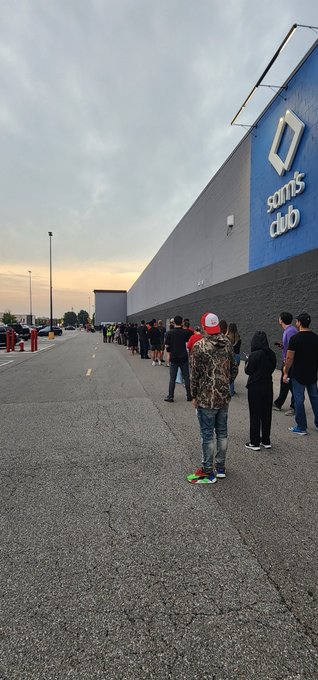 A long line of people stretches along the side of a "Sam's Club" building, waiting outside during what appears to be early morning or dusk, illustrating the patience required for a chance at a limited product drop.