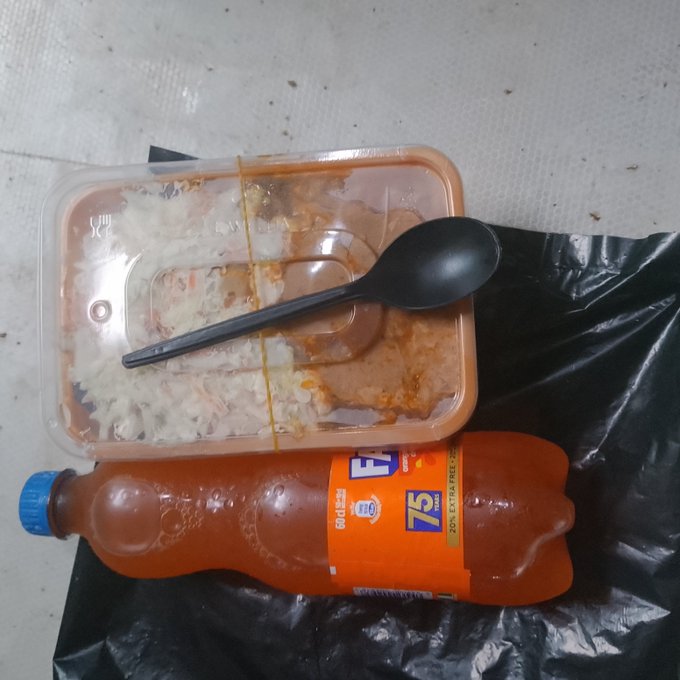 An empty plastic food container with rice remnants and a black spoon sits on a black plastic bag next to a bottle of orange juice labeled '75'.