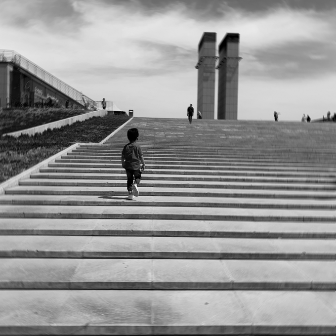 A black-and-white photo of a young boy with a backpack climbing a long flight of stone steps, with towering monolithic structures looming in the distance—evoking the reflective theme of being "a few steps ahead" of those behind us. The image is a singular edition NFT posted on objkt/Teia (Tezos), and any visible text is limited to the artist's caption referencing this single-piece release.