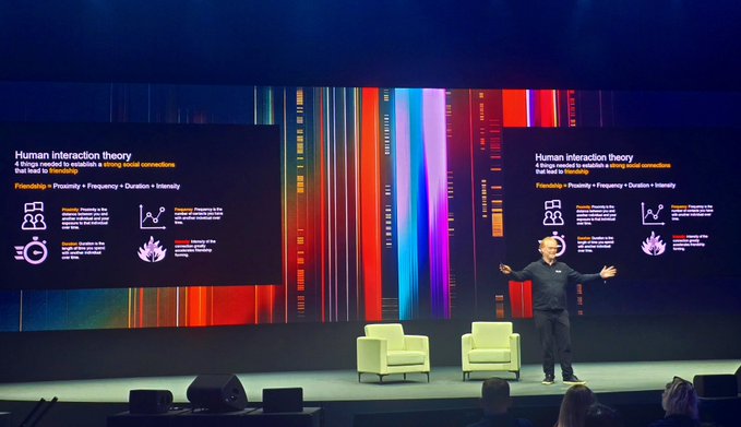 A man giving a presentation on stage in front of two large screens titled "Human interaction theory - If things needed to maintain a strong initial connection that lead to friendship," with a formula of "Friendship = Proximity + Frequency + Duration + Intensity," against a colorful striped backdrop.