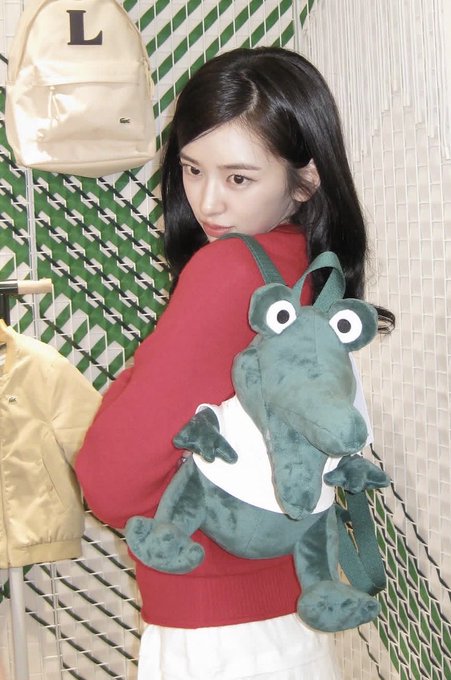 A woman with dark hair poses looking over her shoulder while wearing a red sweater, holding a green plush frog backpack in front of her in a room with patterned green walls.