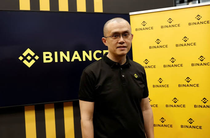 A man wearing glasses and a black polo shirt stands in front of a yellow Binance-branded backdrop with the company's logo.