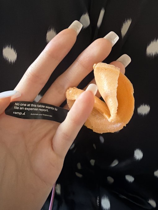 A hand with long white nails holding a fortune cookie, with a fortune slip wrapped around a finger reading "No one at this table wants to file an expense report." with the Ramp logo and "Automate your finance ops."