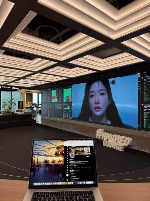 A laptop displaying a Twitter feed sits on a desk in front of a wall-mounted screen showing data analytics, charts, and a woman's face, with a "#HASHED LOUNGE" sign visible. The image reflects the post's mention of being at @hashed_official in Seoul, showcasing a sleek tech-focused workspace likely used for crypto and AI-related work, with @KaitoAI and @triplescosmos referenced in the tweet about ongoing work.