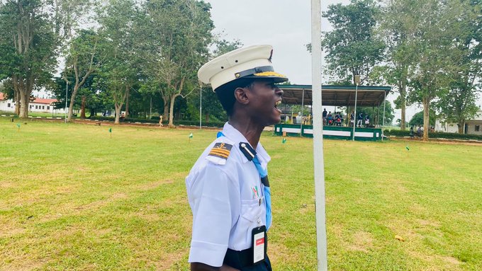 A young man in a white naval-style uniform and cap shouting with his mouth wide open, standing on a grassy field with trees and a covered pavilion in the background.