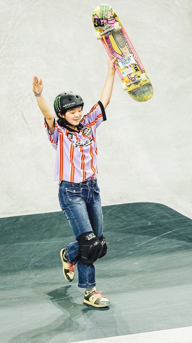 A young skateboarder in a Purple striped jersey and Monster helmet celebrates triumphantly, holding her skateboard aloft in a skate park, matching the post's celebration of Mizuho Hasegawa's record-breaking MVP performance at just 15 years old. The skateboard's colorful graphic deck and her joyful pose capture the excitement of her historic X Games League achievement.