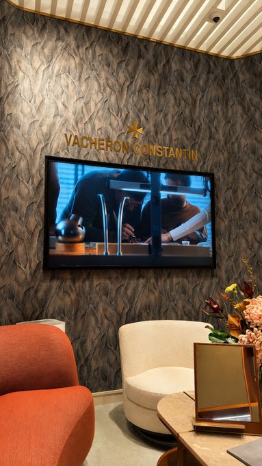A luxurious Vacheron Constantin showroom display features a wall-mounted TV screen showing watchmakers at work, accompanied by elegant orange and cream lounge seating and a fresh floral arrangement. The refined setting reflects the post's excitement about adding a new high-end timepiece to their collection from this prestigious brand.