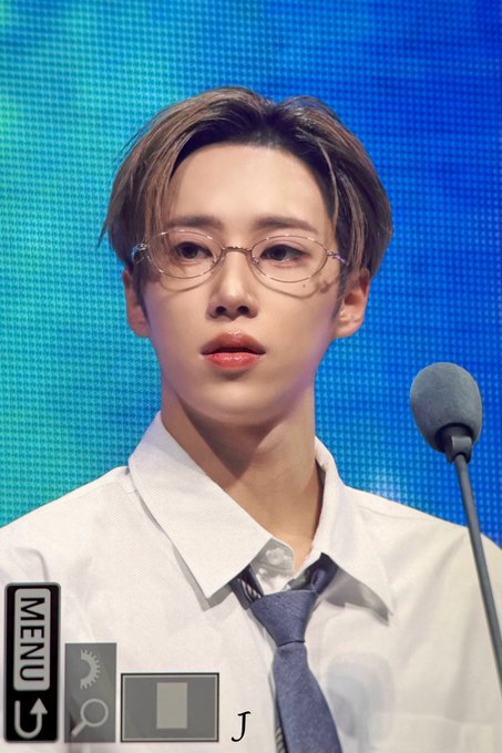 The image features a young man wearing glasses, a white shirt, and a blue tie. He has short hair and looks directly at the camera with an expression of concentration or thoughtfulness. The background appears to be a blue wall with a microphone positioned in front of him. There is also a menu icon visible on the left side of the image.