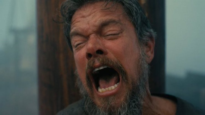 A bearded man with grey hair is captured mid-scream or yawn with his eyes closed and mouth wide open, set against a foggy grey background.
