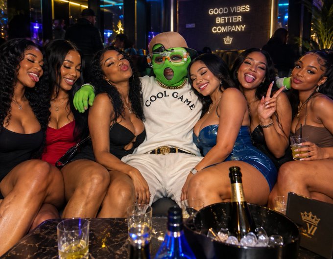 A group photo at a nightclub with a man wearing a green mask, sunglasses, and a 'COOL GAME' sweater posing with six women on his lap, with 'GOOD VIBES BETTER COMPANY' signage visible in the background and an ice bucket with a bottle in the foreground.
