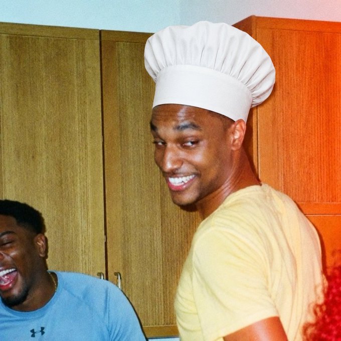 A photograph of NBA player Giannis Antetokounmpo smiling while wearing a white chef's hat, with another man in a blue Under Armour shirt laughing in the background.