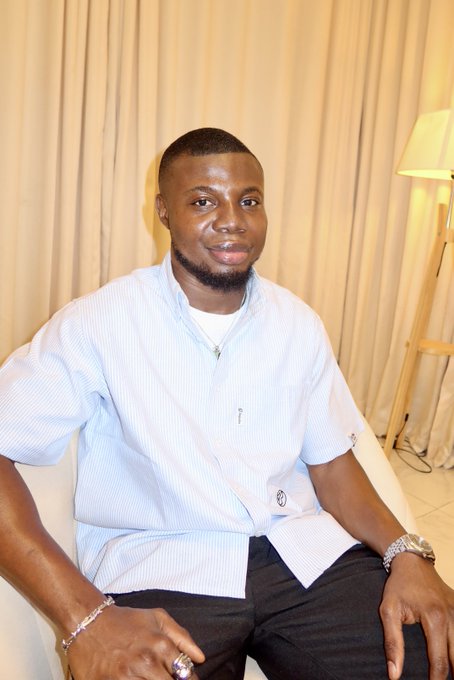 A young man in a light blue striped shirt and dark trousers sits on a white couch with a warm-lit lamp and cream curtains behind him, smiling at the camera in a relaxed portrait. The image complements the introductory post, putting a face to "Ola, 19, AI builder from Nigeria" as he shares his background in building communities, crypto products, and AI agents.