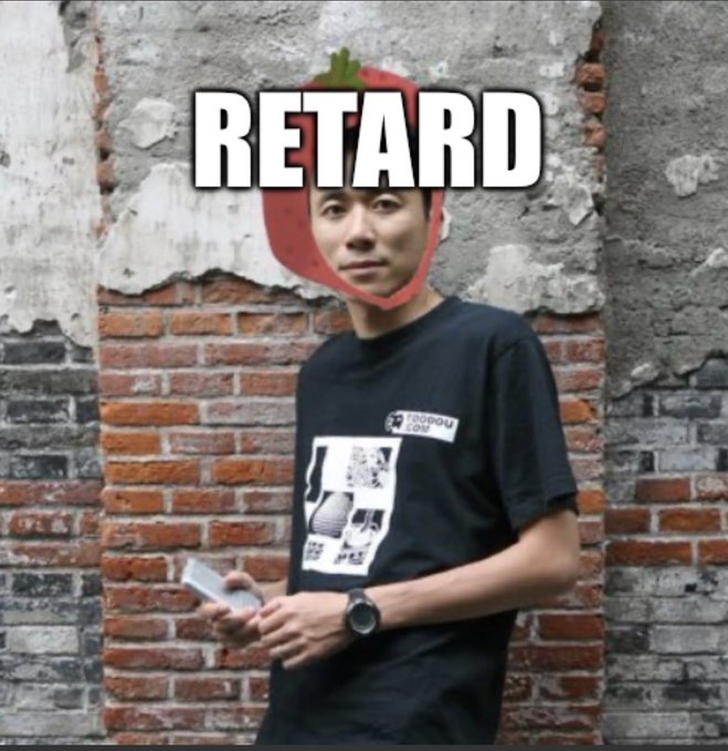 A man in a black graphic t-shirt stands against a brick wall, with a digitally added strawberry on his head and the word "RETARD" overlaid in large white text.