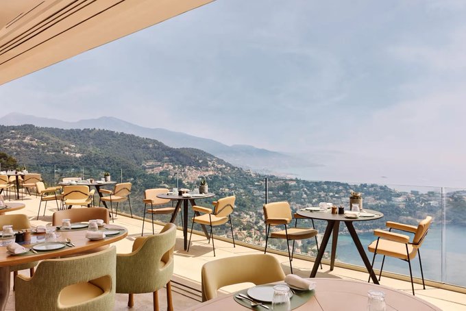 A scenic oceanfront restaurant terrace with empty tables and chairs overlooking mountains and a blue sea under a hazy sky.