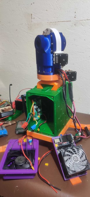 A DIY robotics project featuring a green and orange 3D-printed base with electronic motors, wires, a small fan, and circuit boards, appearing to be an animatronic mechanism.