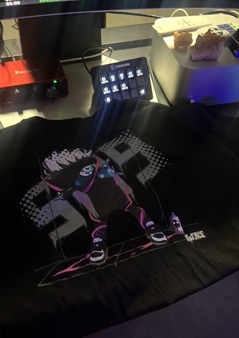 A gaming/streaming desk setup featuring a black mechanical keypad with illuminated keys, a Focusrite audio interface, and a black t-shirt with a pink and white futuristic mechanical design and 'It Gets Worse' text displayed in front of the monitor.
