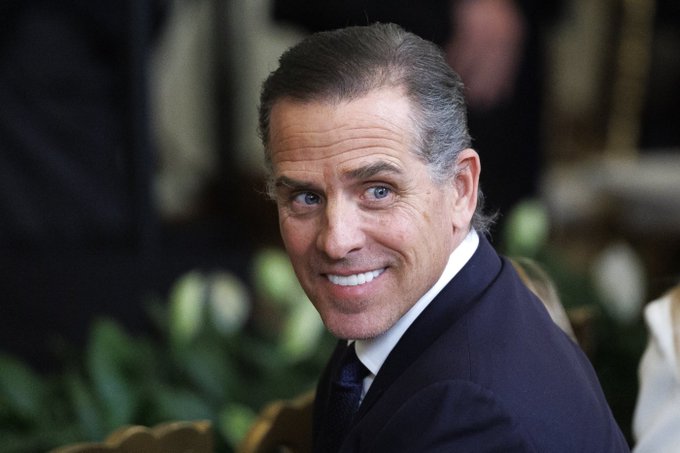 A close-up photograph of a smiling middle-aged man with graying hair wearing a dark suit, looking over his shoulder with a friendly expression.
