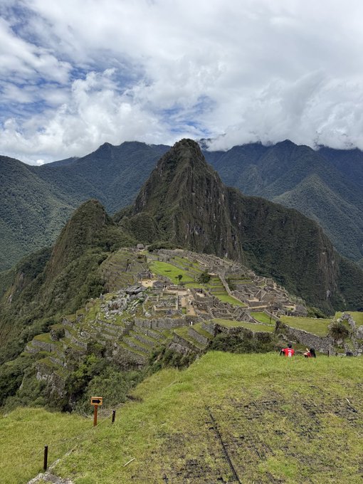 A scenic photograph of the Machu Picchu ruins with its iconic mountain peak in the background, viewed from a grassy hillside.