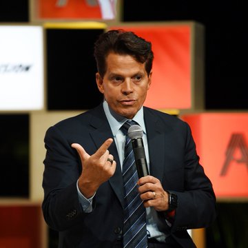 Anthony Scaramucci, dressed in a dark suit and striped tie, speaks into a microphone while gesturing with his hand at what appears to be a panel or conference event. The image relates to the post about his bullish stance on Bitcoin, showing him delivering his market insights.