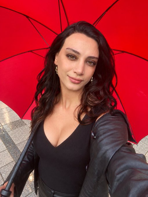 A woman with dark wavy hair poses in front of a bright red umbrella, wearing a black top and leather jacket.