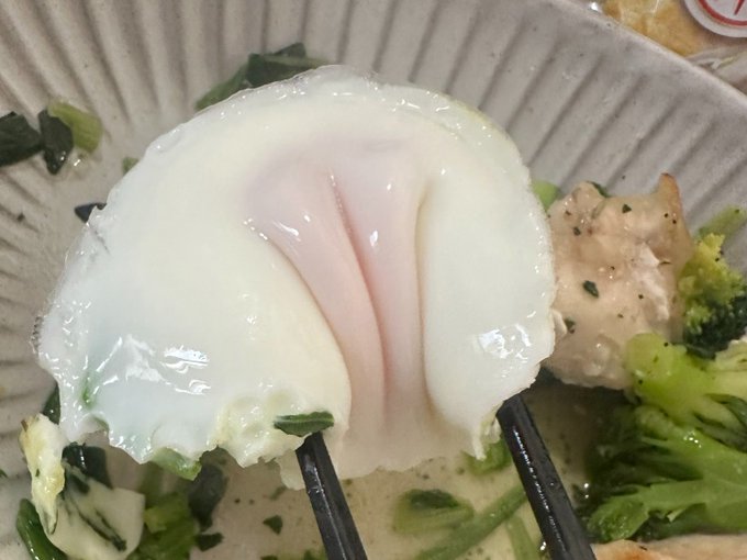 Chopsticks grip a soft poached egg over a bowl containing tofu, chicken, and green vegetables.