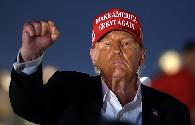 An older man in a dark blazer and white shirt raises his fist while wearing a red "MAKE AMERICA GREAT AGAIN" hat, with a small American flag pin visible on his lapel. The image depicts Donald Trump, the key political figure named in the post as an expected attendee at the White House crypto meeting.