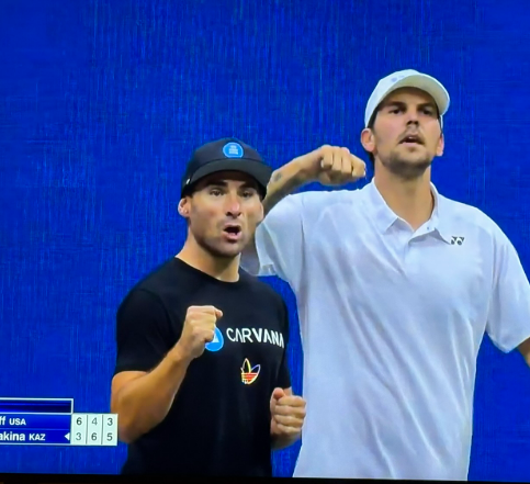 Two tennis players in white shirts and caps pump their fists in celebration during a match; a scoreboard shows 'f usa' and 'akina kaz'.