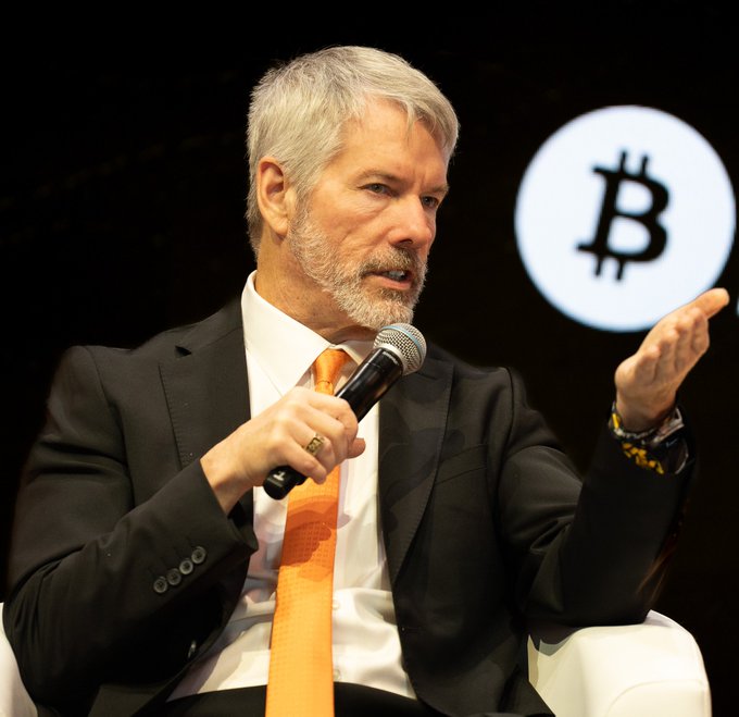 A man in a dark suit and orange tie speaks into a microphone, gesturing with his left hand, while a Bitcoin (₿) logo is displayed in the background—visually reinforcing Michael Saylor's quote defending Bitcoin's foundational principles against rule changes rooted in statist control.