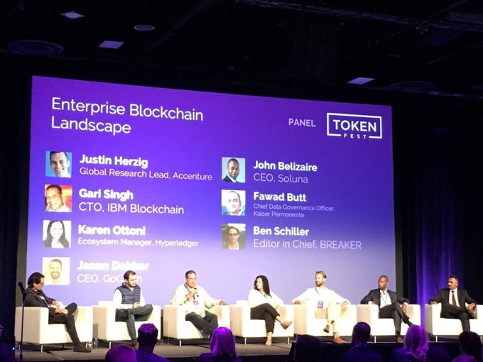 A panel discussion at TOKEN FEST titled 'Enterprise Blockchain Landscape' featuring Justin Herzig (Accenture), John Belizaire (Soluna), Gari Singh (IBM), Fawad Butt (Kaiser Permanente), Karen Ottoni (Hyperledger), and Ben Schiller (BREAKER), with participants seated on stage.