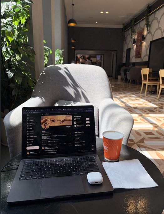 A laptop sits open on a coffee shop table displaying what appears to be a Spotify or music streaming app interface, alongside a coffee cup and AirPods case. The setup reflects the "building from cafe" working environment mentioned in the post, with someone multitasking on various digital projects including development, SEO, and app store optimization.